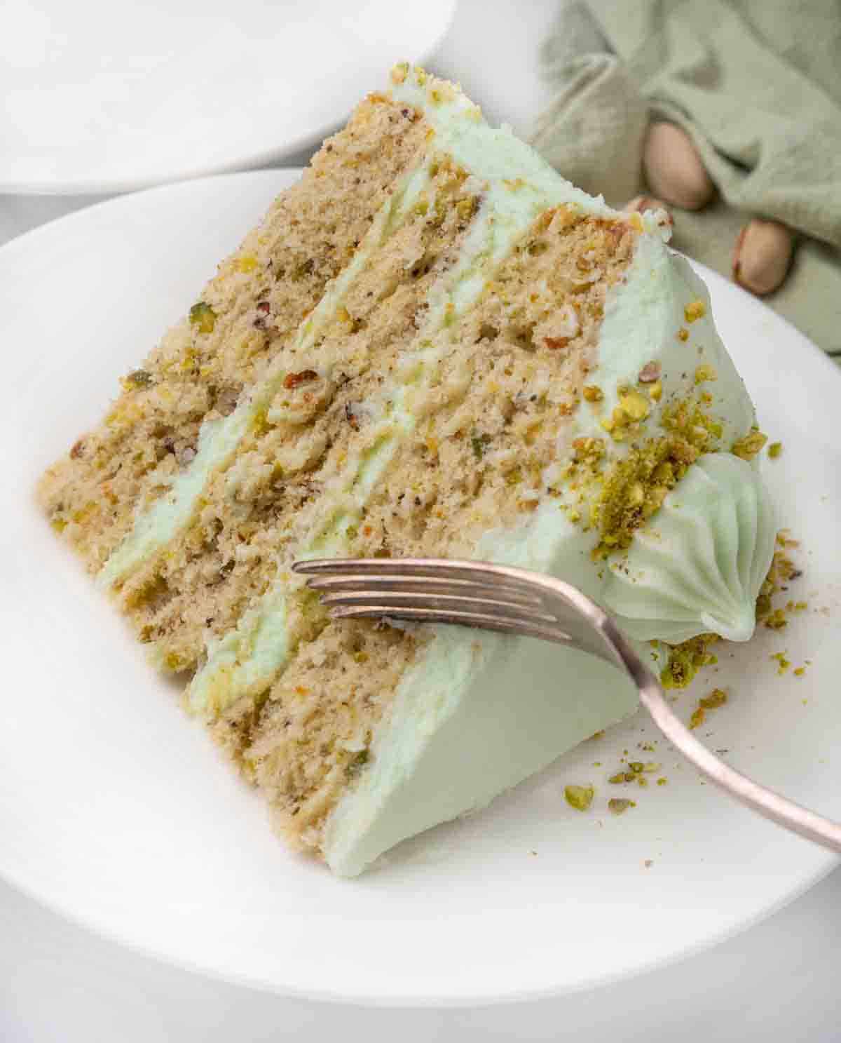 Slice of pistachio cake on a white plate with a fork in the slice.