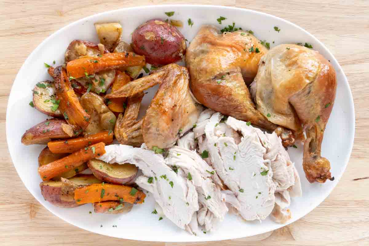 Sliced roasted chicken and whole pieces with vegetables on a white platter.