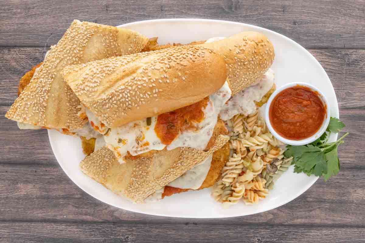 Chicken parmesan sandwich on a white platter with marinara sauce and pasta salad.