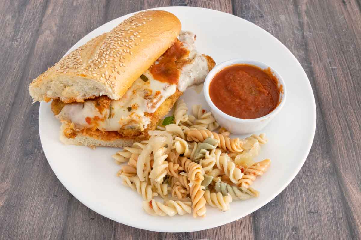 Chicken parmesan sandwich on a white platter with marinara sauce and pasta salad.