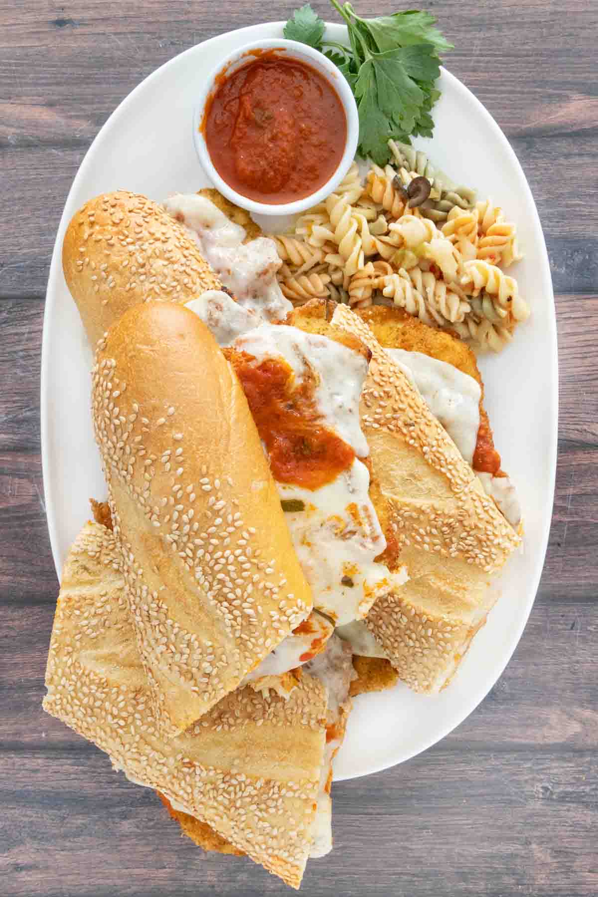 Chicken parmesan sandwich on a white platter with marinara sauce and pasta salad.