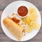 Chicken parmesan sandwich on a white platter with marinara sauce and potato chips.