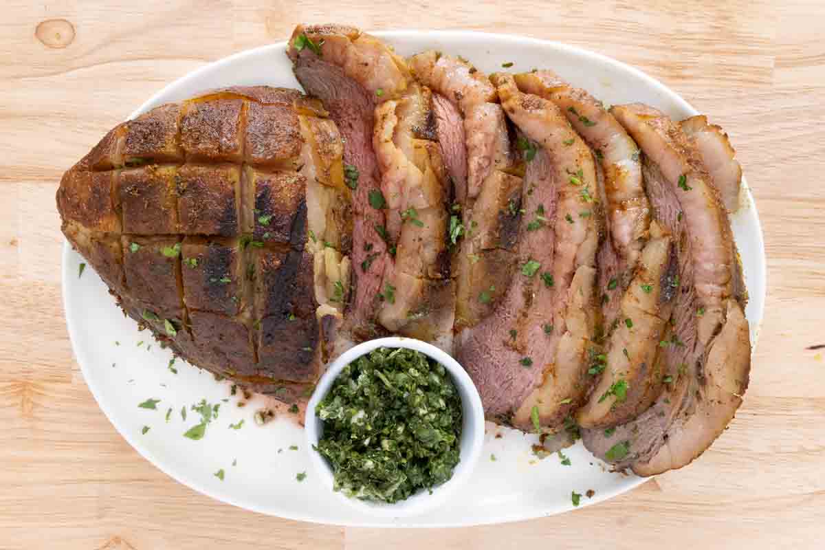 Sliced smoked picanha with a bowl of gremolata on a white platter.