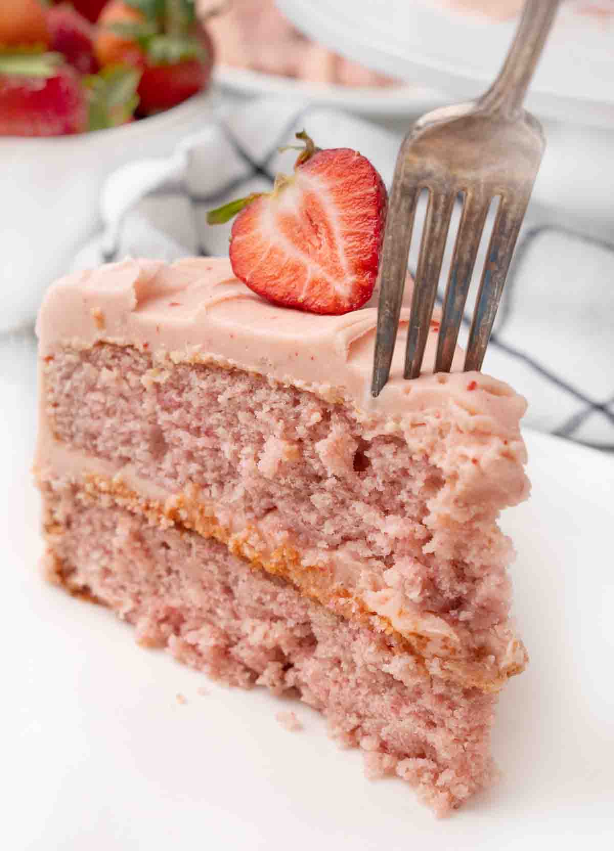 Slice of strawberry cake on a white plate with a fork going into the slice.