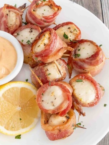 Scallops wrapped in bacon on a white plate with dipping sauce and half lemon.