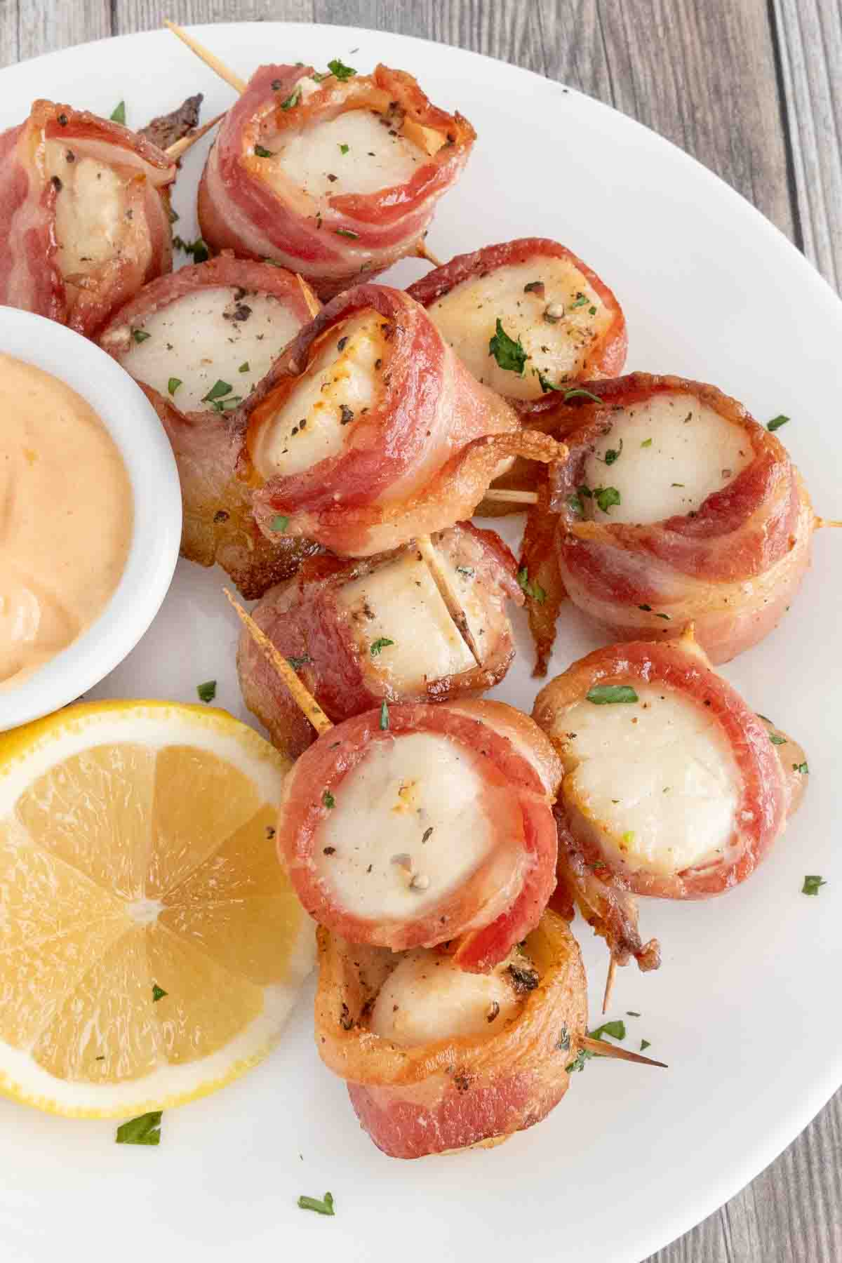 Scallops wrapped in bacon on a white plate with dipping sauce and half lemon.