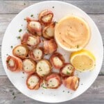 Scallops wrapped in bacon on a white plate with dipping sauce and half lemon.