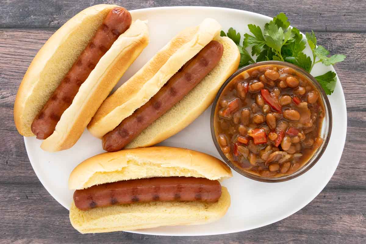 Baked beans in a glass bowl with hot dogs on a white plate.