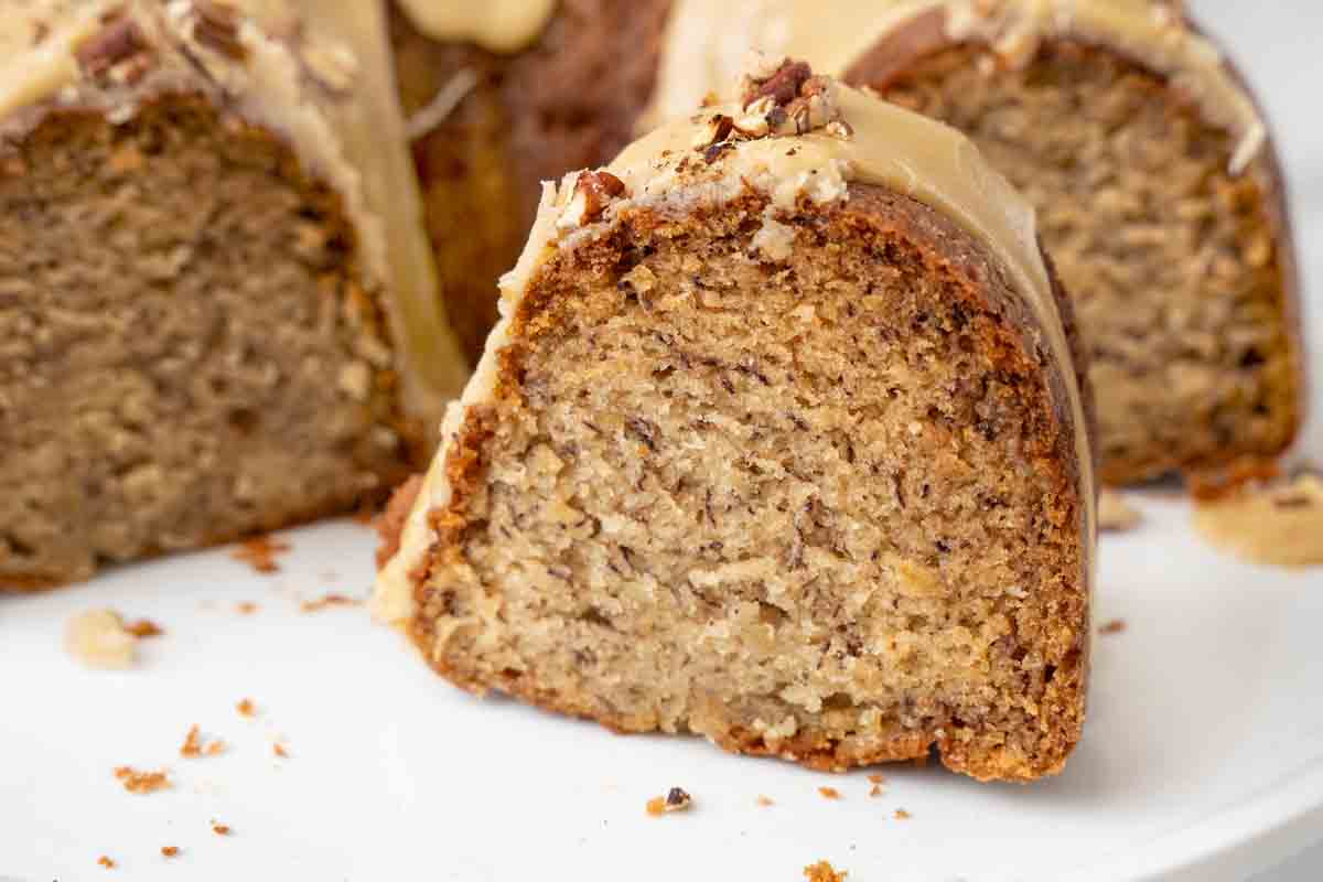 Slice of banana bundt cake on a white platter in from of the rest of the cake.