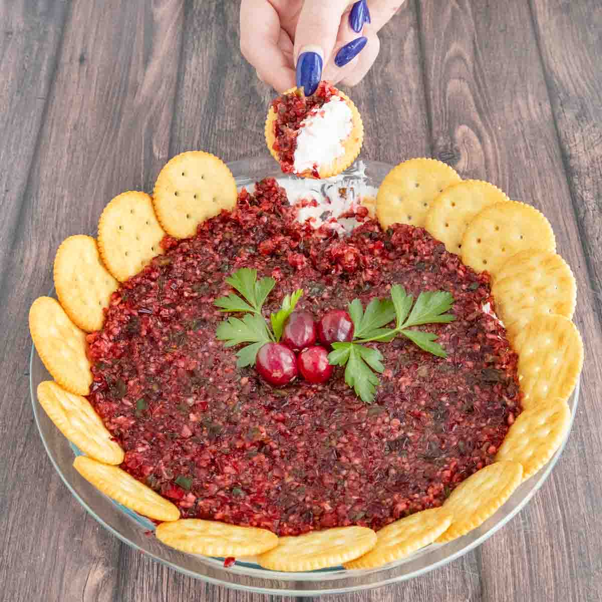 Cranberry Jalapeno dip with a ring of crackers and a hand dipping a cracker.